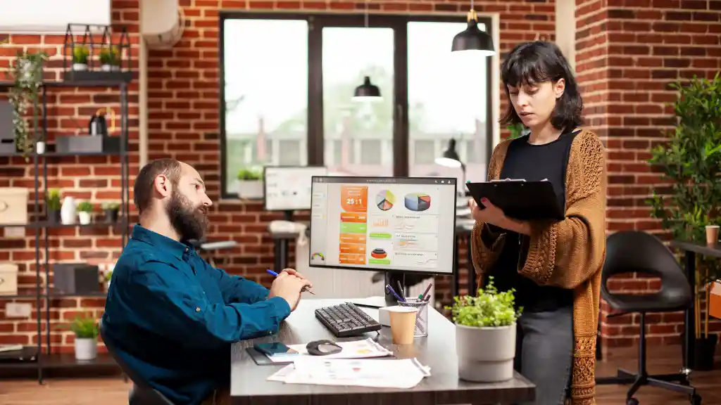 Two people discussing business data shown on a computer monitor in an office.